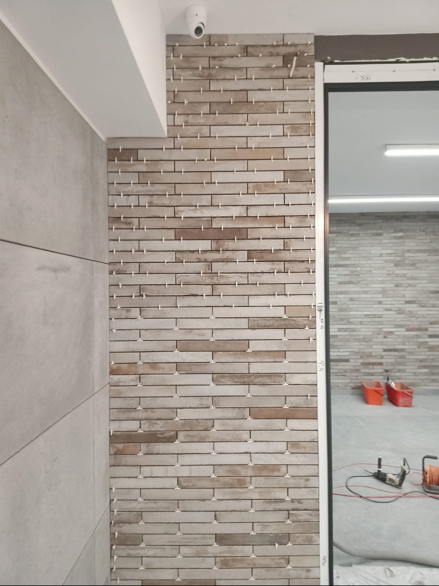 Układanie beżowo-brązowych płytek imitujących cegłę na ścianie, z widocznymi krzyżykami dystansowymi. W tle widoczne narzędzia i wiaderka.