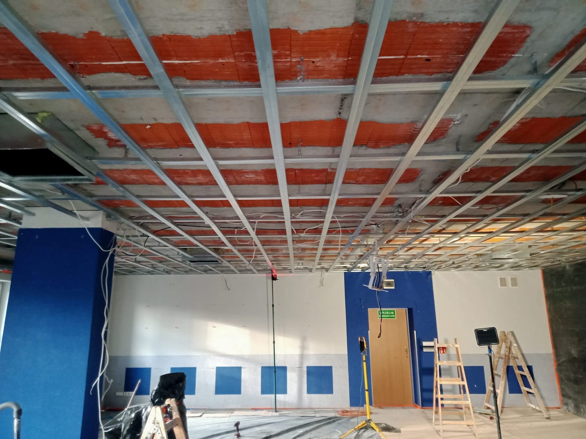 Montaż metalowej konstrukcji sufitu podwieszanego w pomieszczeniu z niebieskimi akcentami na ścianach. Widoczne drabiny i narzędzia, sugerujące etap prac wykończeniowych.