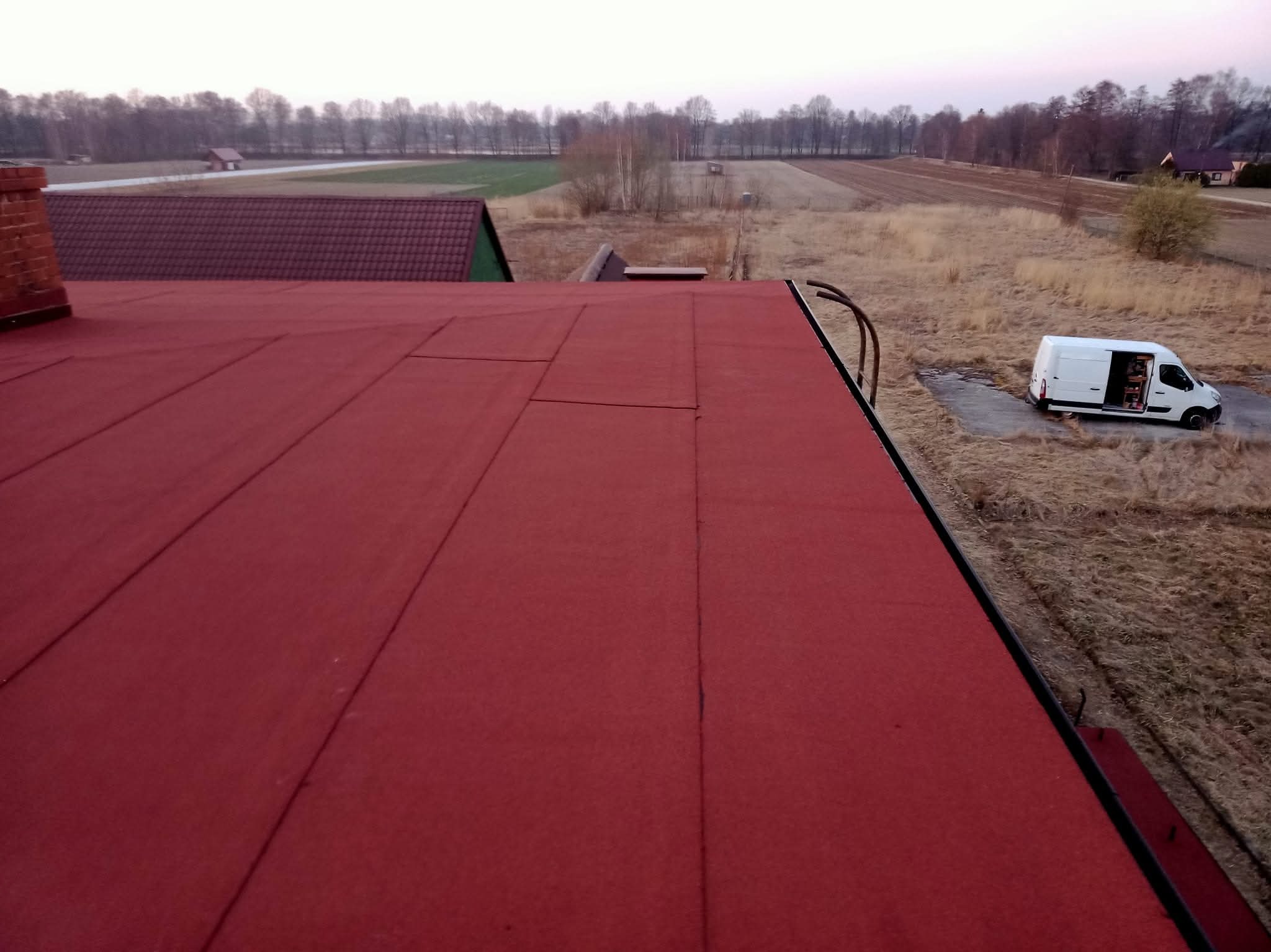 Czerwony dach z papy na tle krajobrazu z polami i lasem, widoczny biały samochód dostawczy. Ujęcie z góry, prezentujące wykończoną powierzchnię dachu.