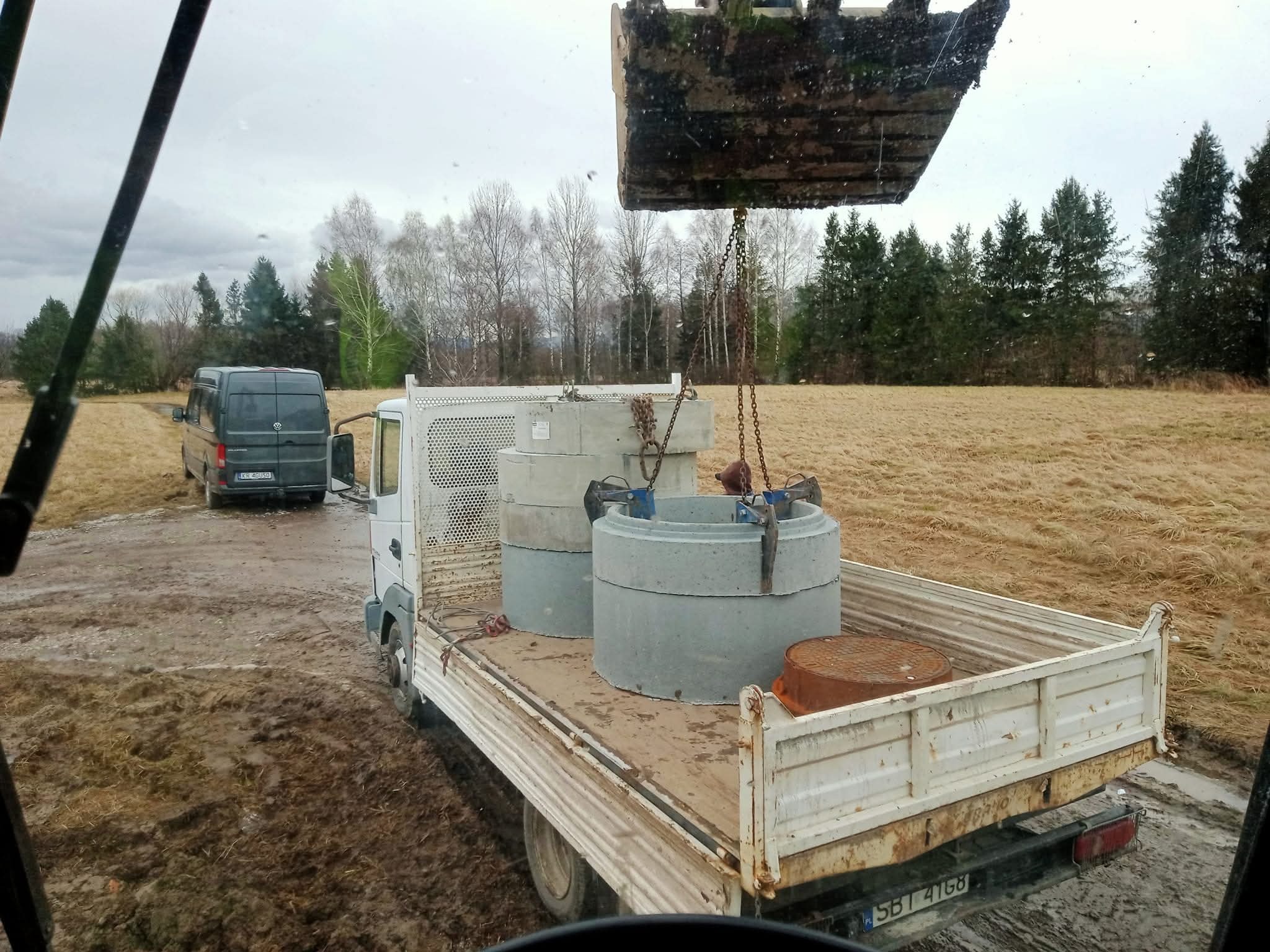 Transport betonowych kręgów studziennych na pace białej ciężarówki, z widoczną łyżką koparki w tle, na błotnistym terenie. Widoczny również szary bus.