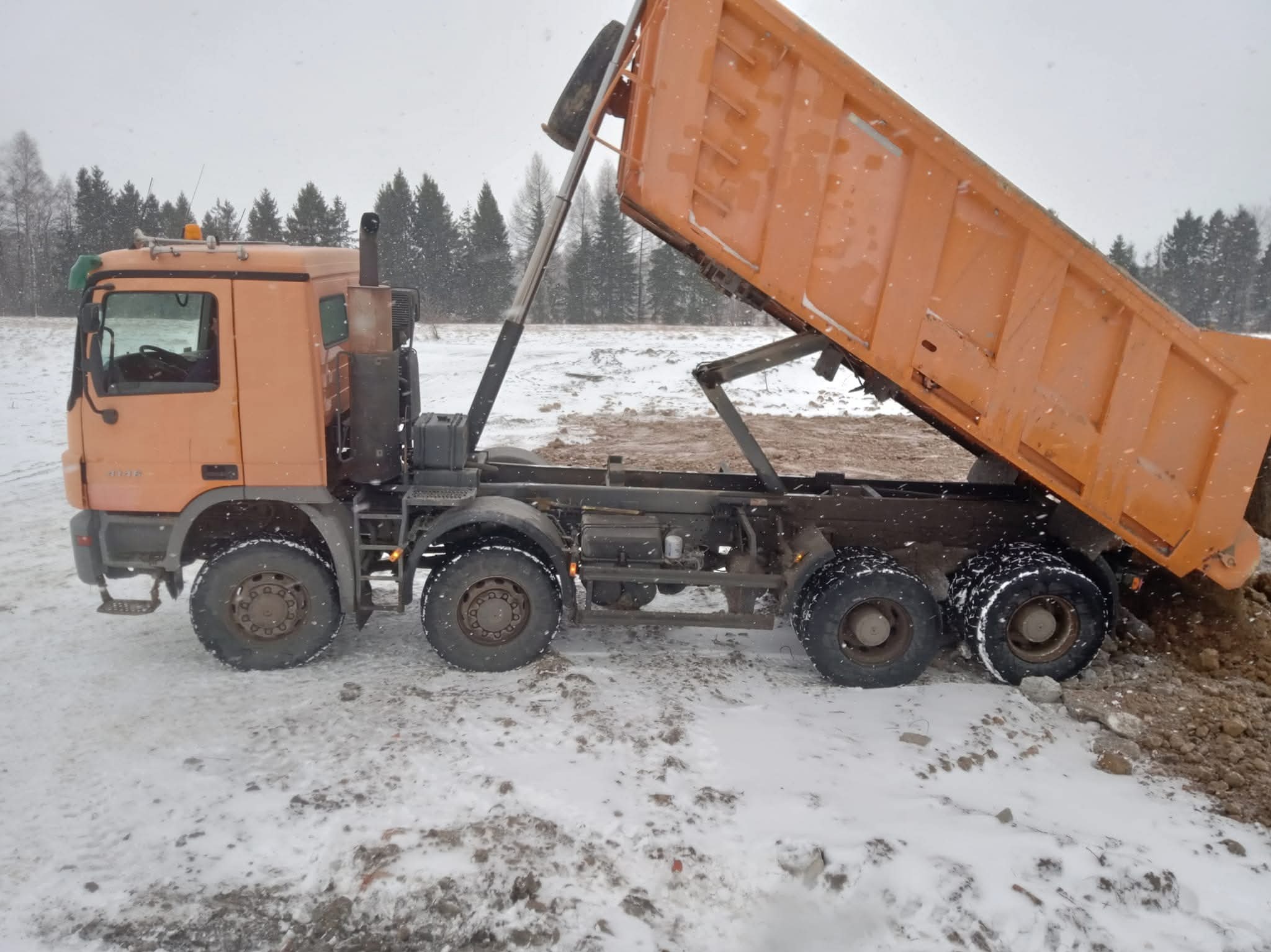 Pomarańczowa wywrotka w trakcie rozładunku ziemi na zasypanym śniegiem terenie, w tle las iglasty i padający śnieg.