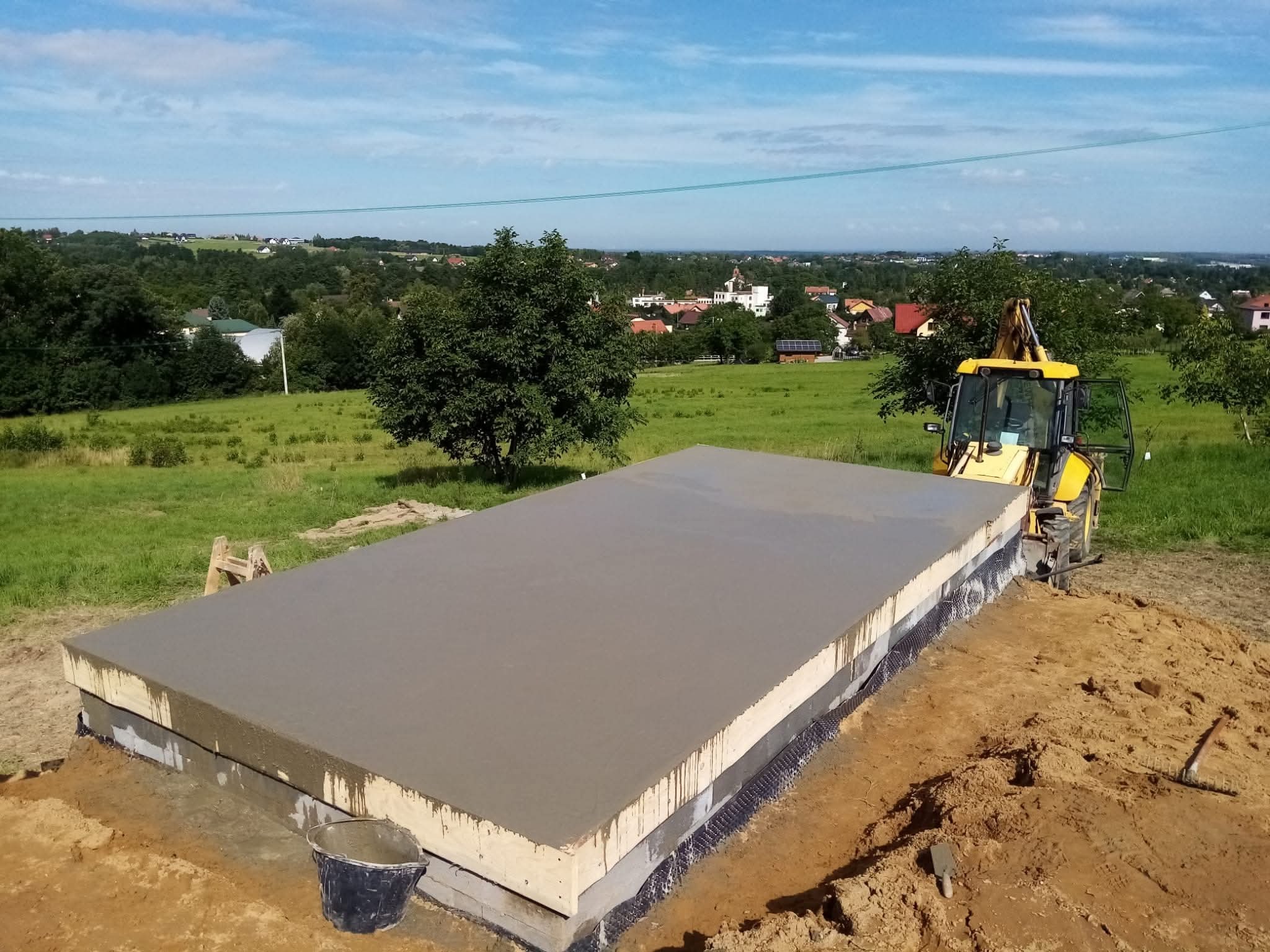 Wylany fundament pod budynek z widoczną izolacją, na tle zielonego krajobrazu i błękitnego nieba. Żółta koparka stoi obok, a w pobliżu leżą narzędzia.