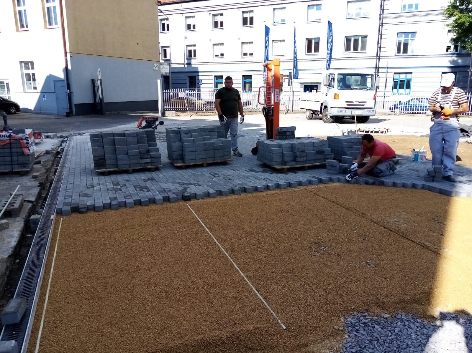 Układanie kostki brukowej na podsypce piaskowej. Pracownicy w trakcie pracy, palety z kostką w tle. Widoczny podnośnik do palet i budynki w tle.