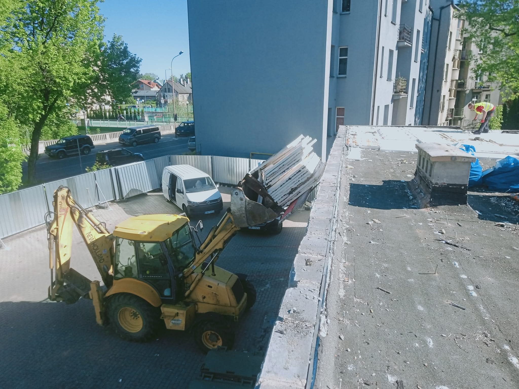 Żółta koparko-ładowarka New Holland zbiera gruz z dachu budynku, w tle biały bus i fragment budynku mieszkalnego. Prace rozbiórkowe w toku.