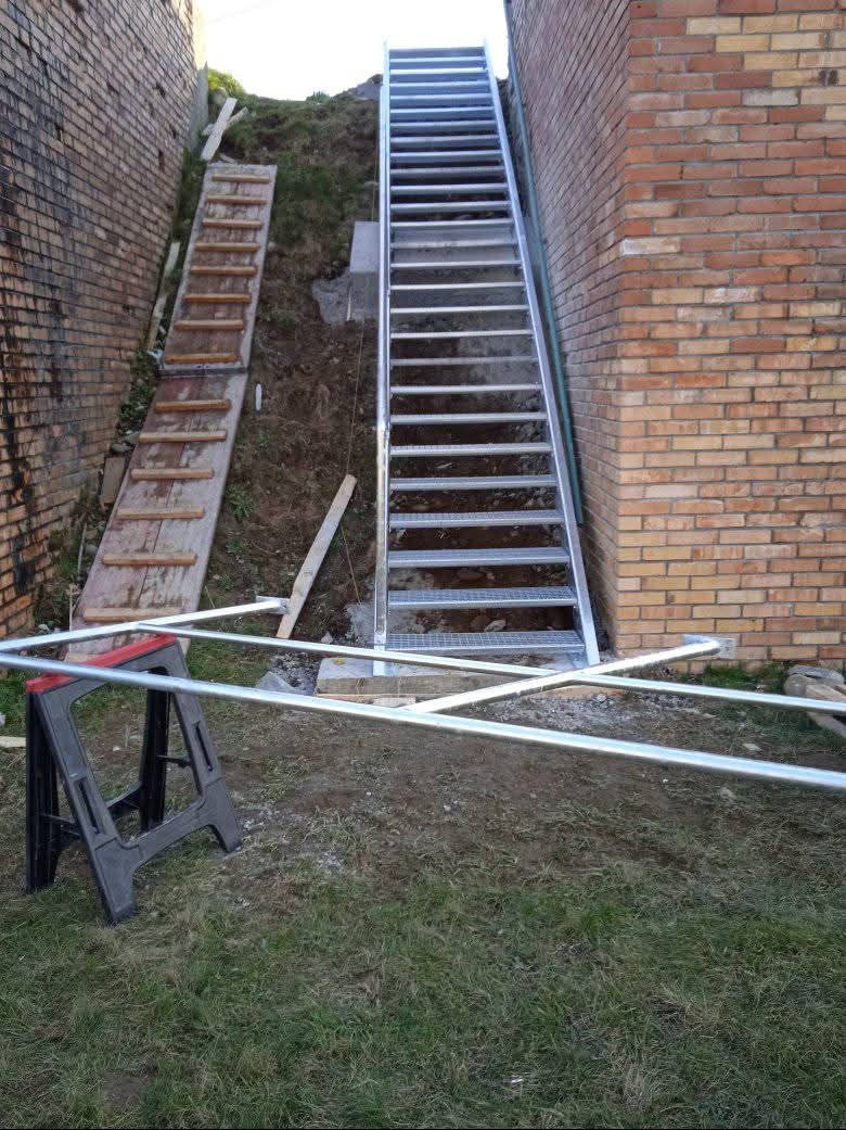 Montaż metalowych schodów zewnętrznych na zboczu, widoczne tymczasowe schody drewniane, ceglany mur po bokach, narzędzia pracy na trawie.