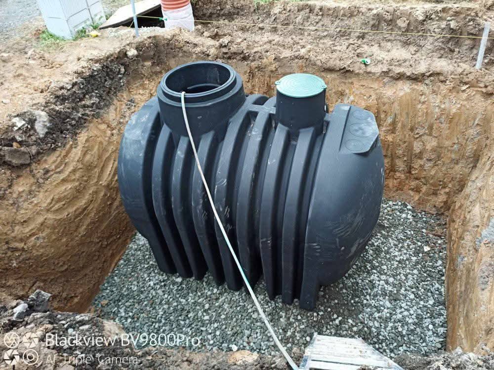 Czarny zbiornik na szambo umieszczony w wykopie, obsypany drobnym żwirem. Widoczne ściany wykopu z ziemi. Zielona pokrywa na zbiorniku.