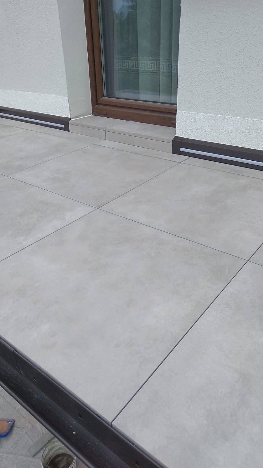 Jasnoszara, kwadratowa podłoga z płytek na zewnątrz budynku, z widocznym oknem i fragmentem ściany. Widoczne fugi i listwy przypodłogowe.