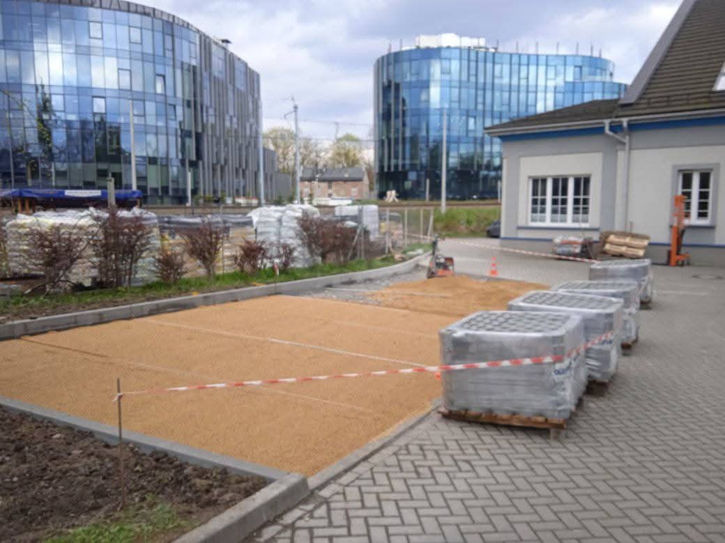 Przygotowany teren pod budowę parkingu z kostki brukowej, widoczne palety z kostką, w tle budynki biurowe o nowoczesnej architekturze.