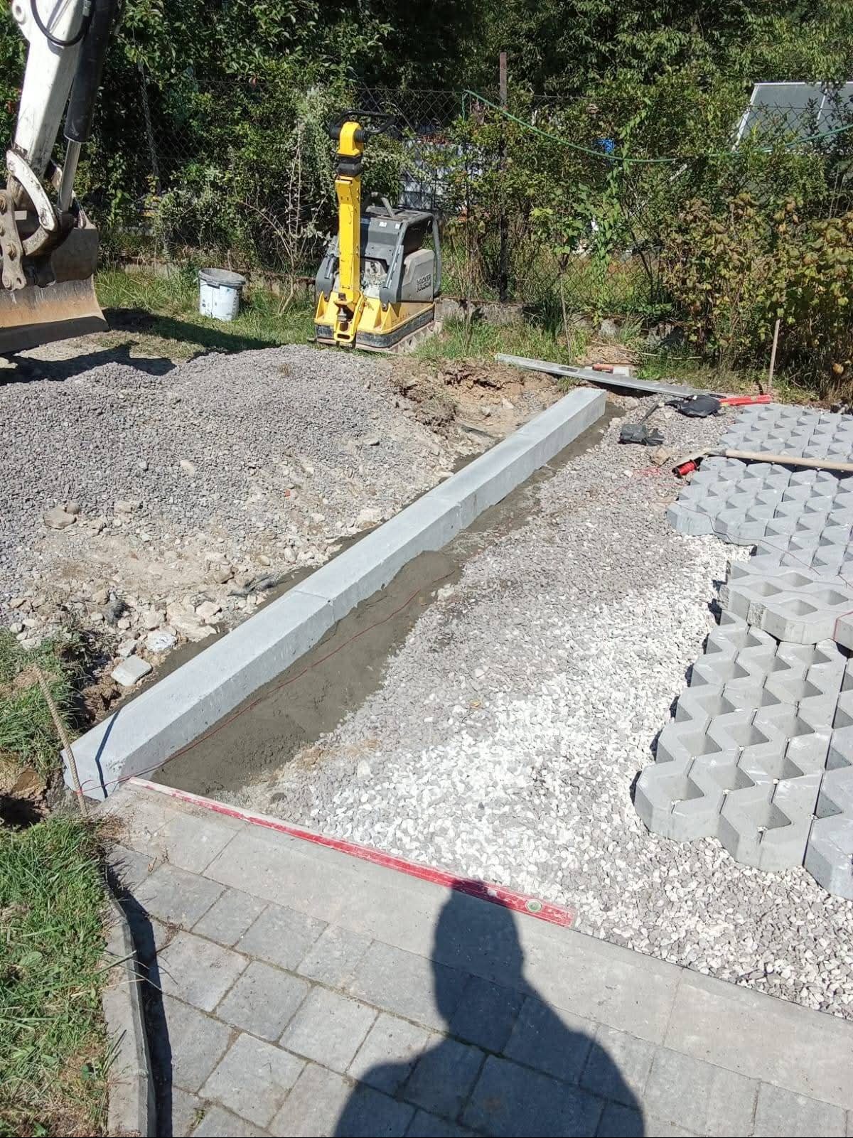 Układanie obrzeży betonowych i kostki ażurowej na podsypce żwirowej. W tle ubijarka i fragment koparki. Widoczny cień osoby fotografującej.