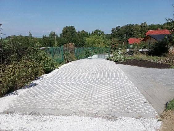 Ukończony podjazd z ażurowych płyt betonowych, widok z poziomu gruntu, zieleń w tle, dom z czerwonym dachem w oddali. Jasne, słoneczne oświetlenie.