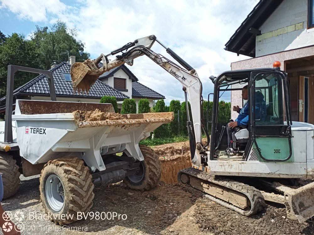Biała mini koparka Bobcat nabiera ziemię do wywrotki Terex na tle domu w trakcie budowy. Operator w kabinie. Wykop pod fundamenty lub instalacje.