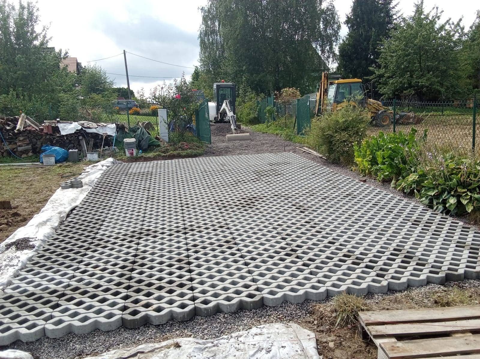 Ułożona nawierzchnia z ażurowych płyt betonowych na podwórku, z widocznymi maszynami budowlanymi w tle. Widok na częściowo ukończony podjazd.
