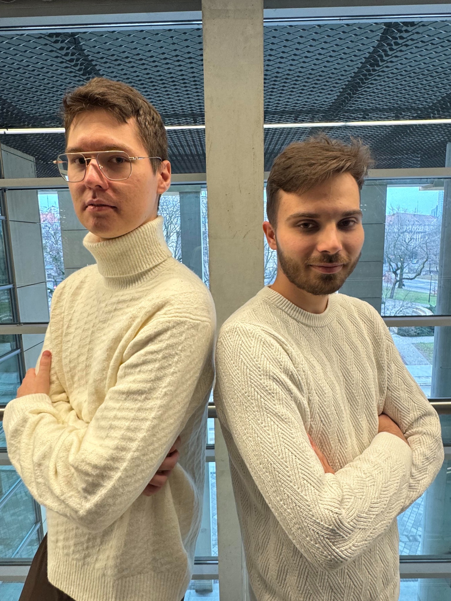 Portret dwóch młodych mężczyzn w jasnych swetrach, stojących plecami do siebie na tle nowoczesnego budynku z dużymi oknami i metalową konstrukcją sufitu.