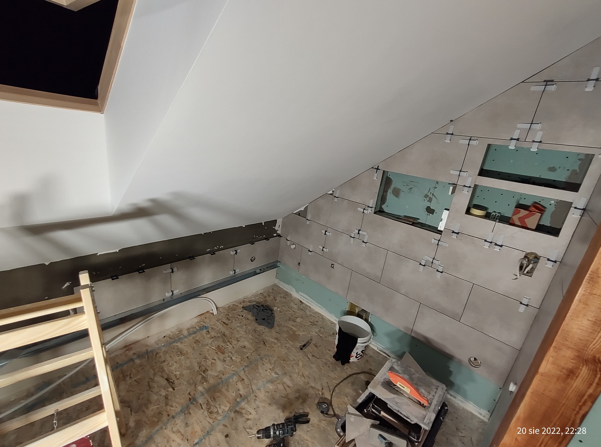 Płytki ceramiczne w trakcie montażu na ścianie poddasza, widoczne krzyżyki dystansowe, narzędzia glazurnicze i drabina. Ukośny sufit i okno dachowe.