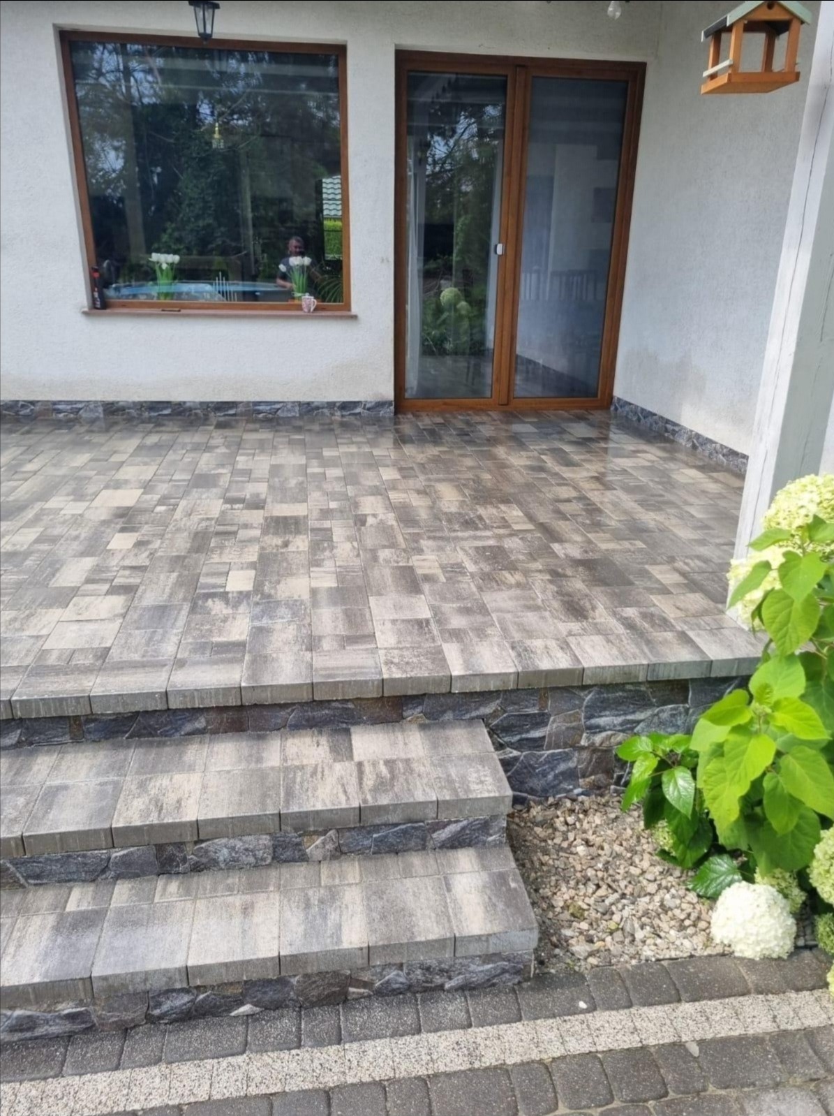 Ułożona kostka brukowa na tarasie z schodami, z widoczną roślinnością i elementami elewacji budynku. Kostka w odcieniach szarości, ułożona w geometryczny wzór.