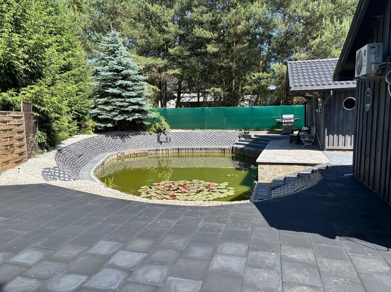 Oczko wodne z liliami wodnymi otoczone kostką brukową i ozdobnym murem z bloczków, w tle iglaki i budynek z szarą elewacją.