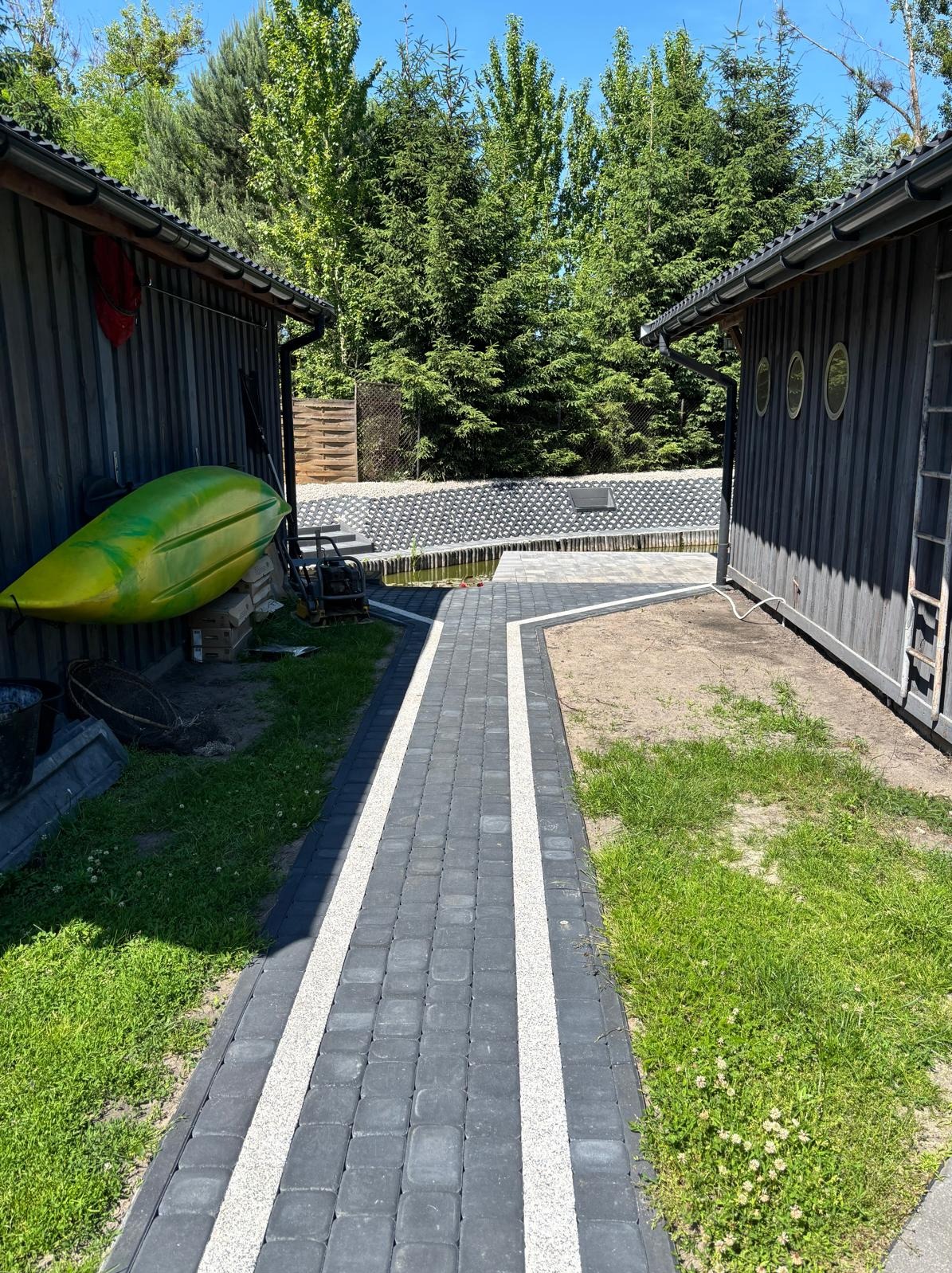 Ścieżka z ciemnej kostki brukowej z białymi pasami, prowadząca między dwoma budynkami z szarego drewna, w tle widoczny staw i zieleń.