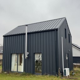 Eliz-Klima Wiktoria Leśko - Nowoczesny dom z szarej blachy trapezowej z klimatyzatorem na zewnątrz. Widoczny komin i okna z białymi firankami. Pochmurne niebo w tle.