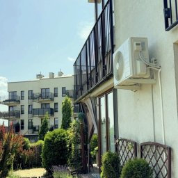 Eliz-Klima Wiktoria Leśko - Biały klimatyzator na elewacji budynku z balkonem, widok z ogrodu z ozdobnymi krzewami i ścieżką. W tle budynek mieszkalny.