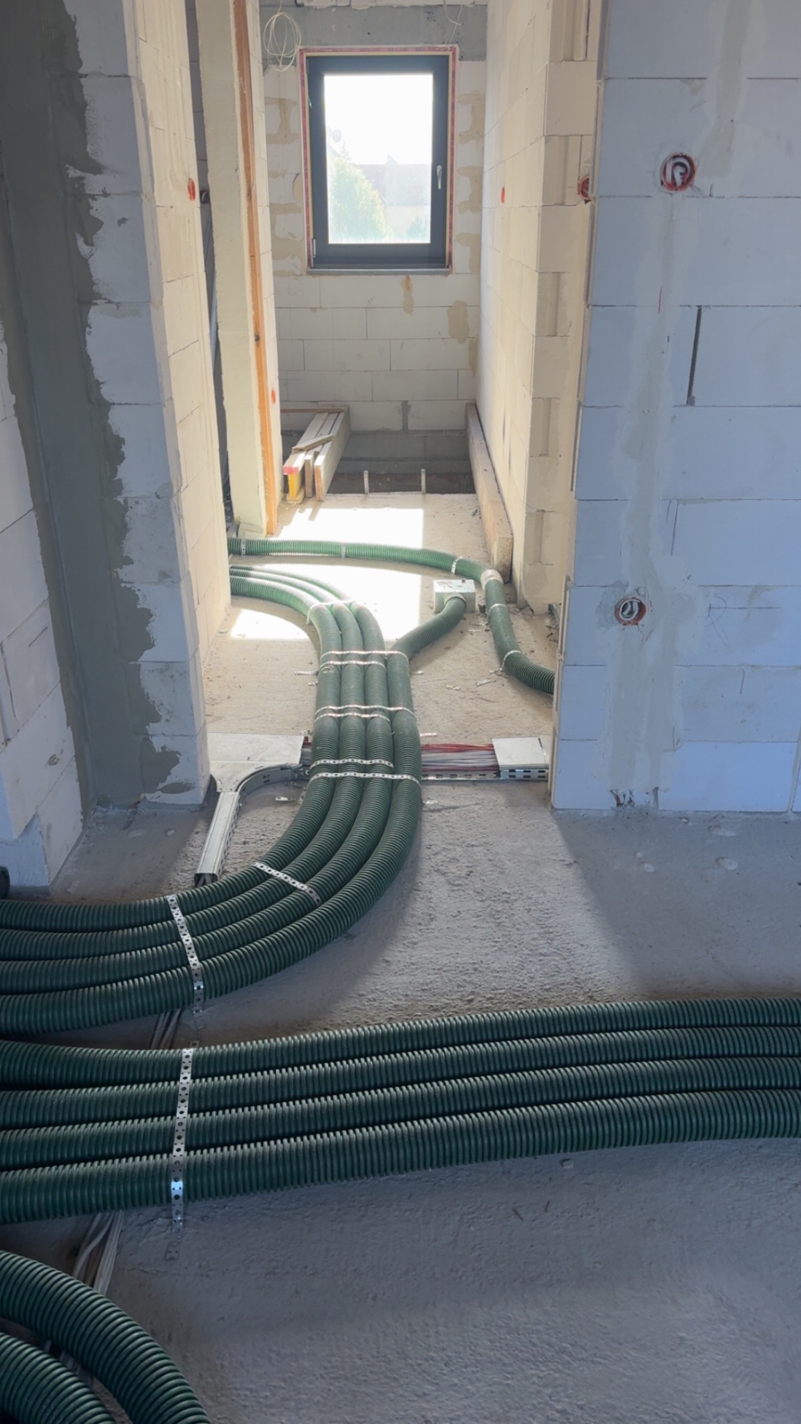 Zielone rury wentylacyjne ułożone równolegle na podłodze w surowym wnętrzu budynku z oknem w tle. Widoczne mocowania rur i fragmenty instalacji elektrycznej.