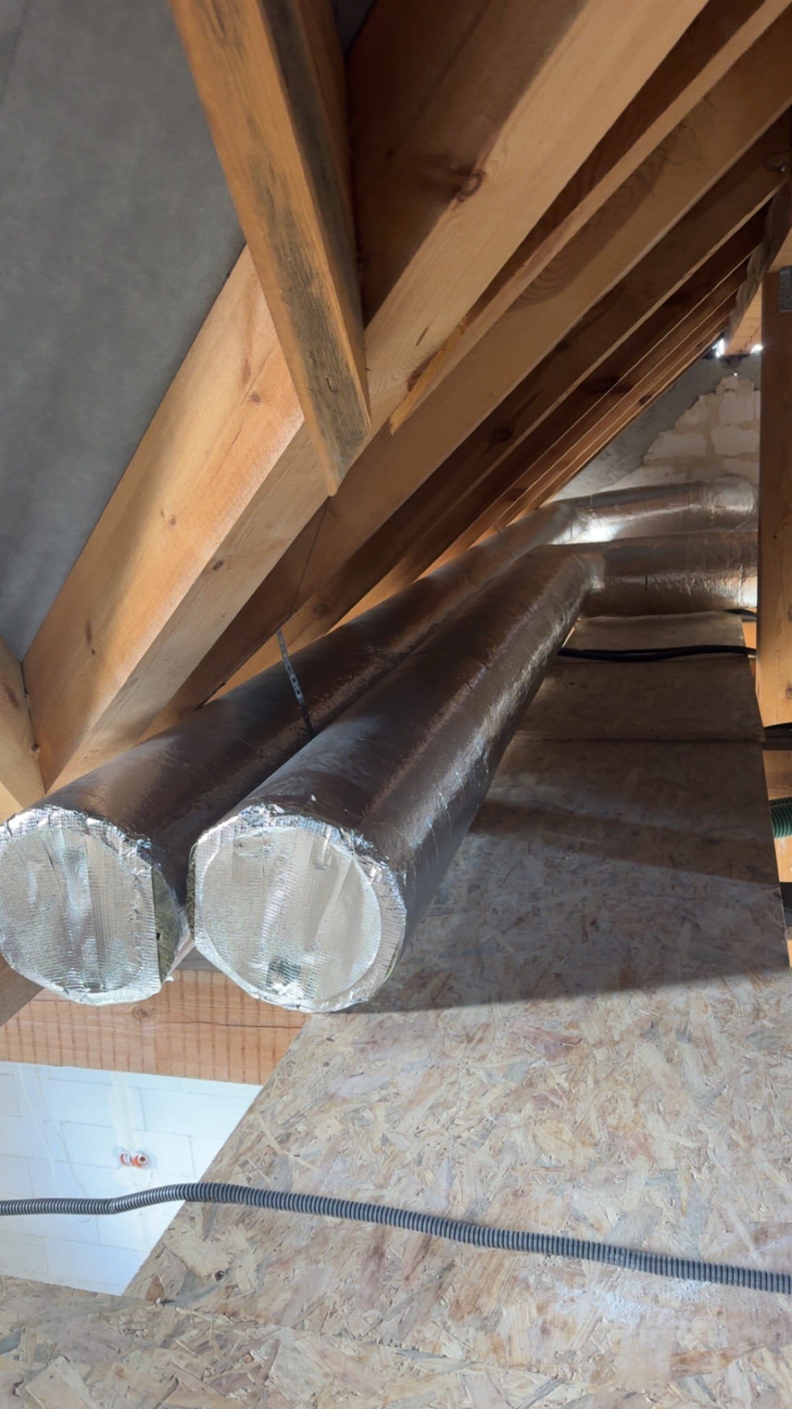 Dwa srebrne, izolowane przewody wentylacyjne zamontowane pod drewnianą konstrukcją dachu na poddaszu, widoczne elementy instalacji elektrycznej.