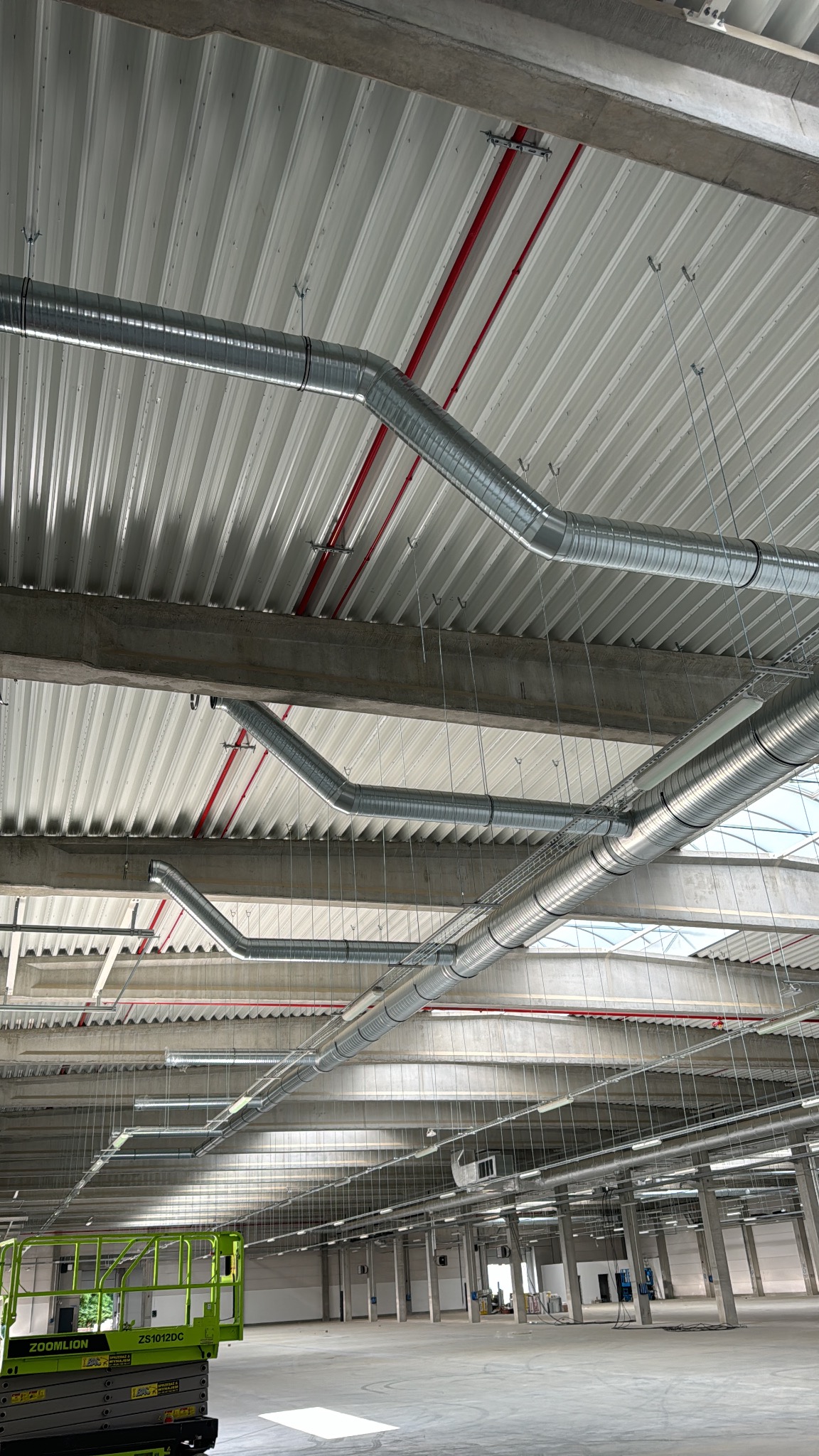 Srebrne rury wentylacyjne pod sufitem hali magazynowej, widoczne czerwone rury i podnośnik nożycowy Zoomlion. Oświetlenie sufitowe i betonowe filary w tle.