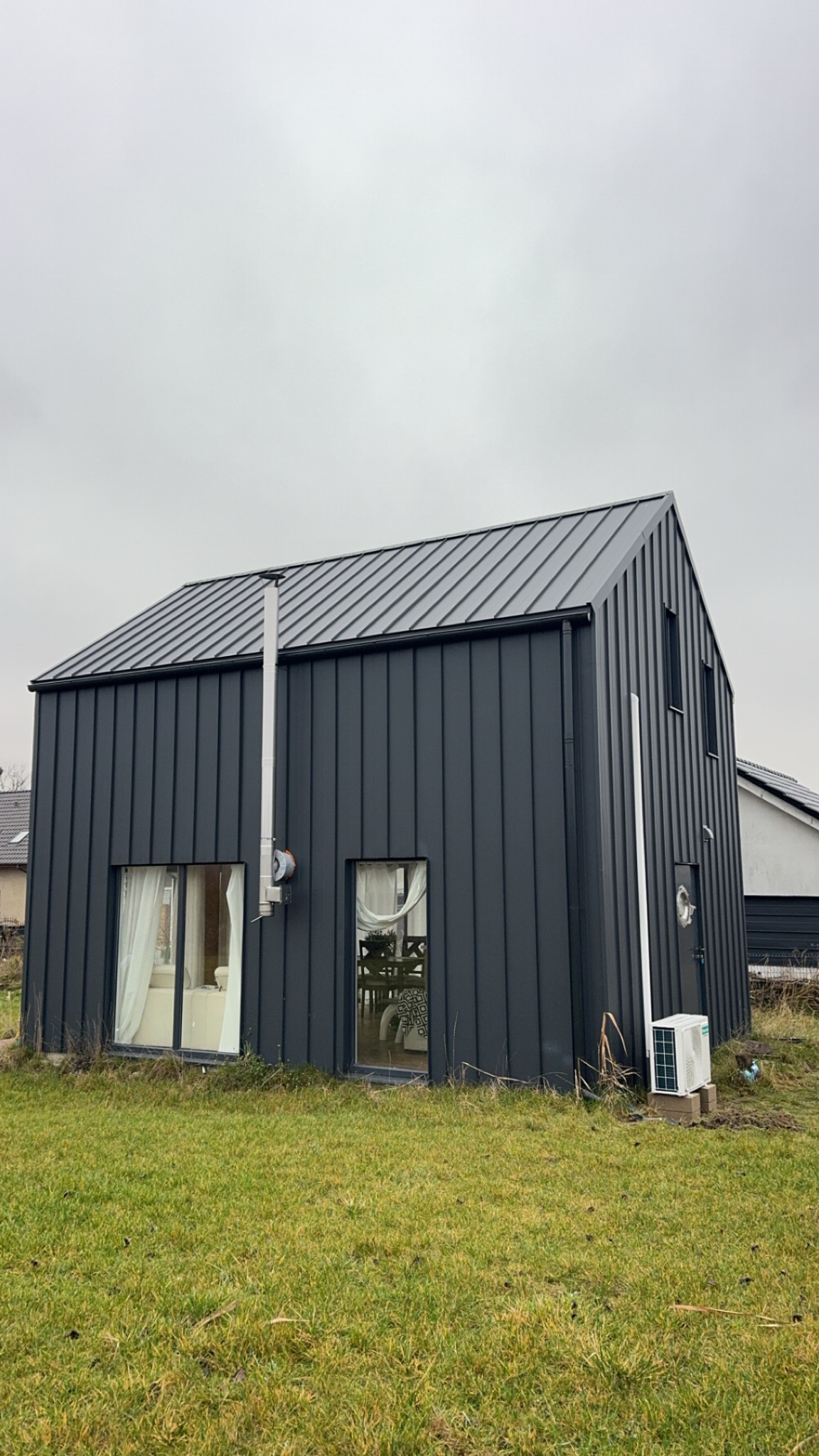 Nowoczesny dom z szarej blachy trapezowej z klimatyzatorem na zewnątrz. Widoczny komin i okna z białymi firankami. Pochmurne niebo w tle.