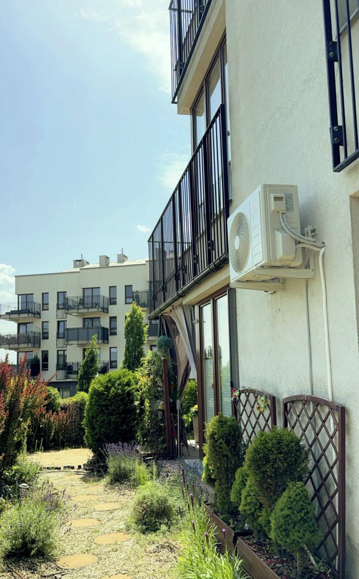 Biały klimatyzator na elewacji budynku z balkonem, widok z ogrodu z ozdobnymi krzewami i ścieżką. W tle budynek mieszkalny.