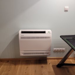 COLD-RES Dominik Markowski - Biały klimatyzator przypodłogowy Sinclair na ścianie w pokoju, widoczne gniazdka elektryczne i fragment biurka gamingowego. Oświetlenie naturalne.