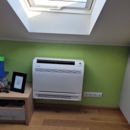 COLD-RES Dominik Markowski - Biały klimatyzator Sinclair na zielonej ścianie pod oknem dachowym. Obok stolik z zabawkami i ramką. Widoczne gniazdka elektryczne i drewniana podłoga.