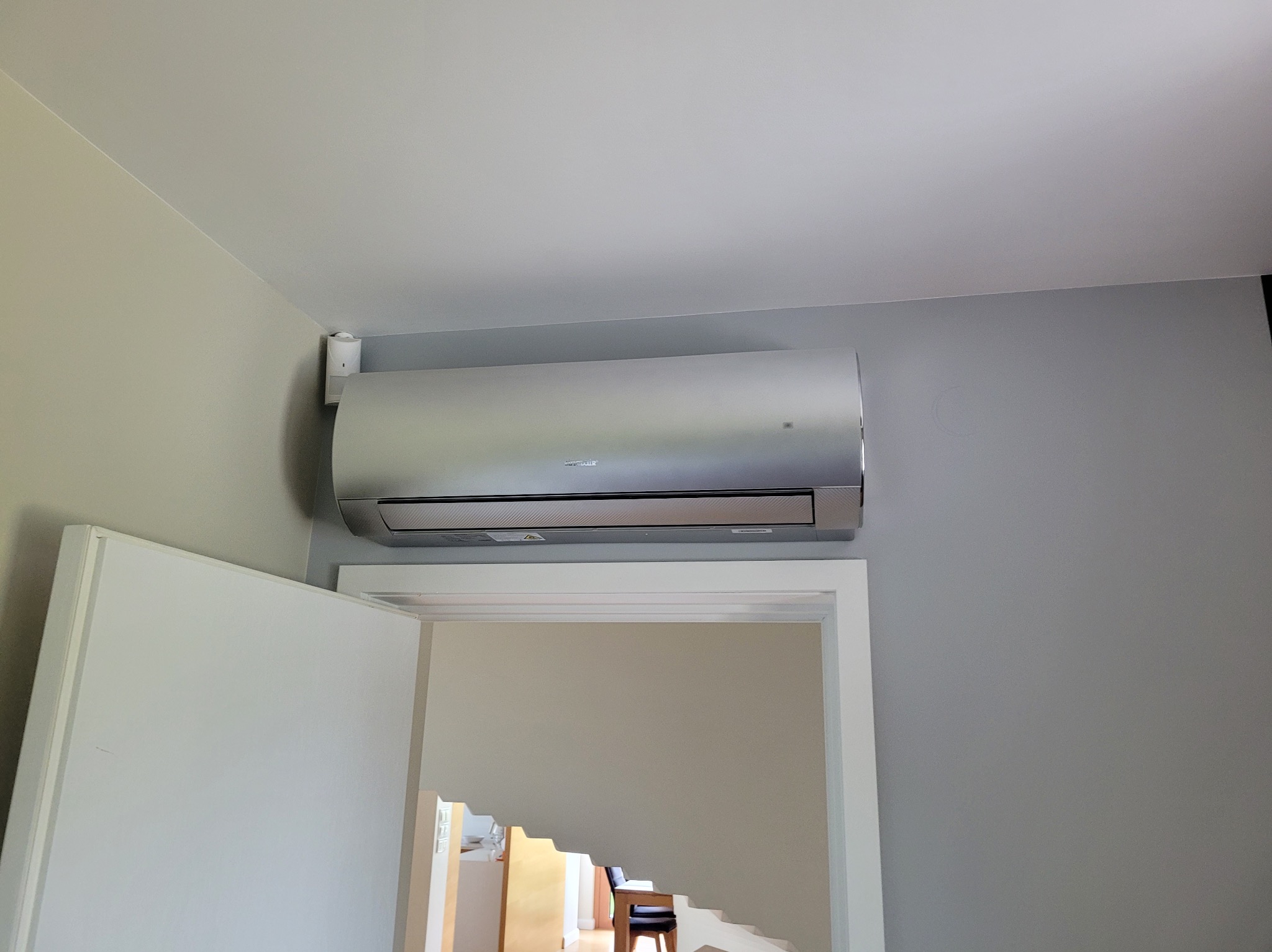 Srebrny klimatyzator ścienny zamontowany nad białymi drzwiami w szarym pokoju. Widoczny czujnik ruchu. Wnętrze domu z schodami w tle.