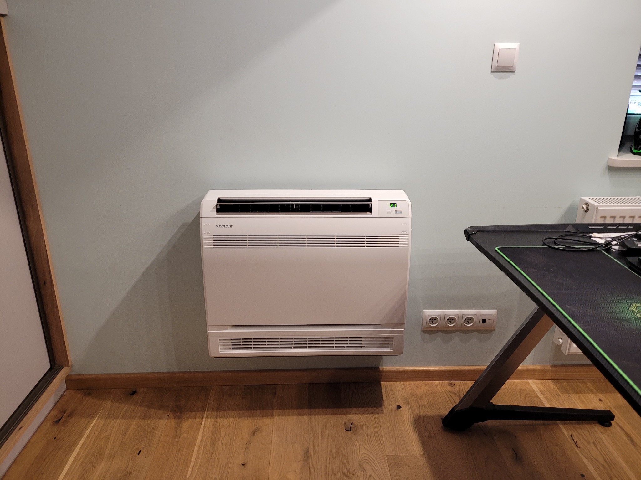 Biały klimatyzator przypodłogowy Sinclair na ścianie w pokoju, widoczne gniazdka elektryczne i fragment biurka gamingowego. Oświetlenie naturalne.