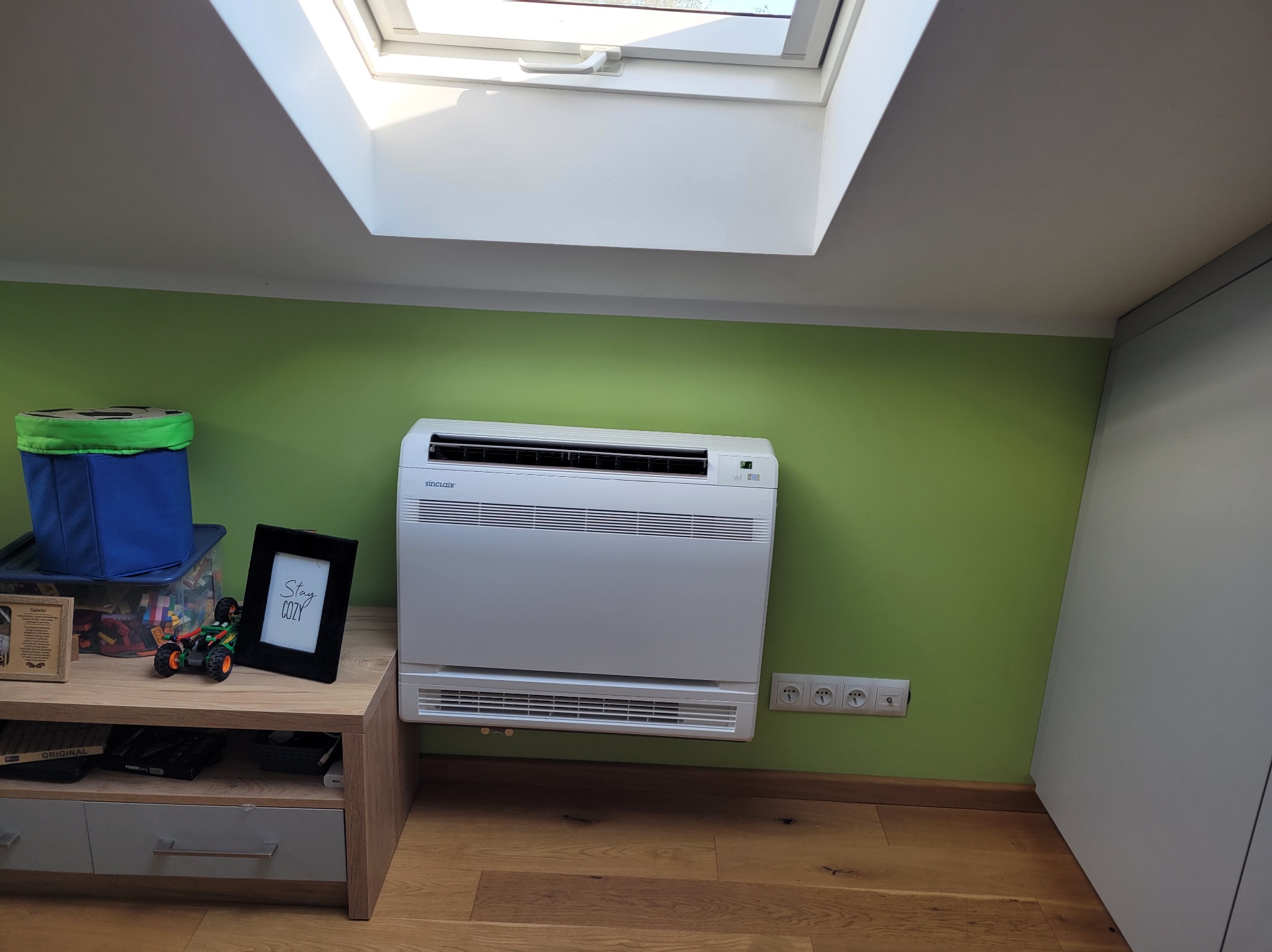 Biały klimatyzator Sinclair na zielonej ścianie pod oknem dachowym. Obok stolik z zabawkami i ramką. Widoczne gniazdka elektryczne i drewniana podłoga.