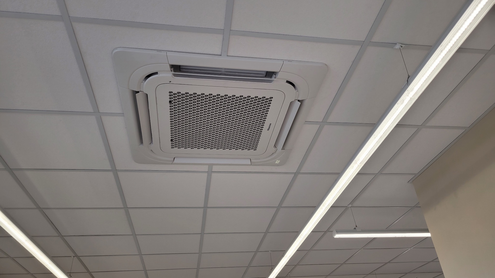 Kasetonowa klimatyzacja Hisense zamontowana w suficie podwieszanym, widok z dołu, oświetlenie liniowe LED w tle. Biała kolorystyka urządzenia i sufitu.