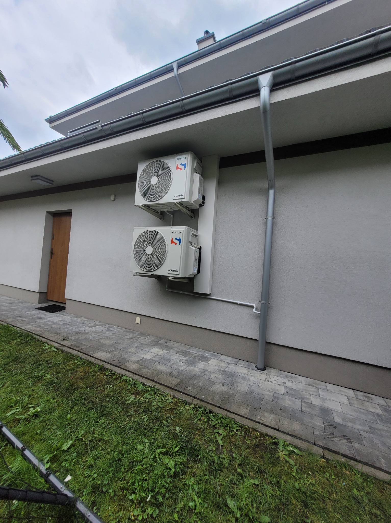 Dwa białe klimatyzatory Sinclair zamontowane na elewacji domu, z widocznymi rurami odprowadzającymi skropliny i elementami instalacji zewnętrznej.