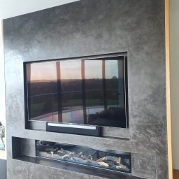 Artur Wojtysiak - Ściana z dekoracyjnym tynkiem w odcieniach szarości, z wnęką na telewizor i kominek elektryczny. Widoczne drewniane elementy wykończeniowe.