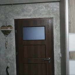 Artur Wojtysiak - Wykończone wnętrze: dekoracyjna ściana z efektem betonu, ciemne drzwi z szybą i szafa przesuwna z drewnopodobnym wzorem i białymi panelami.