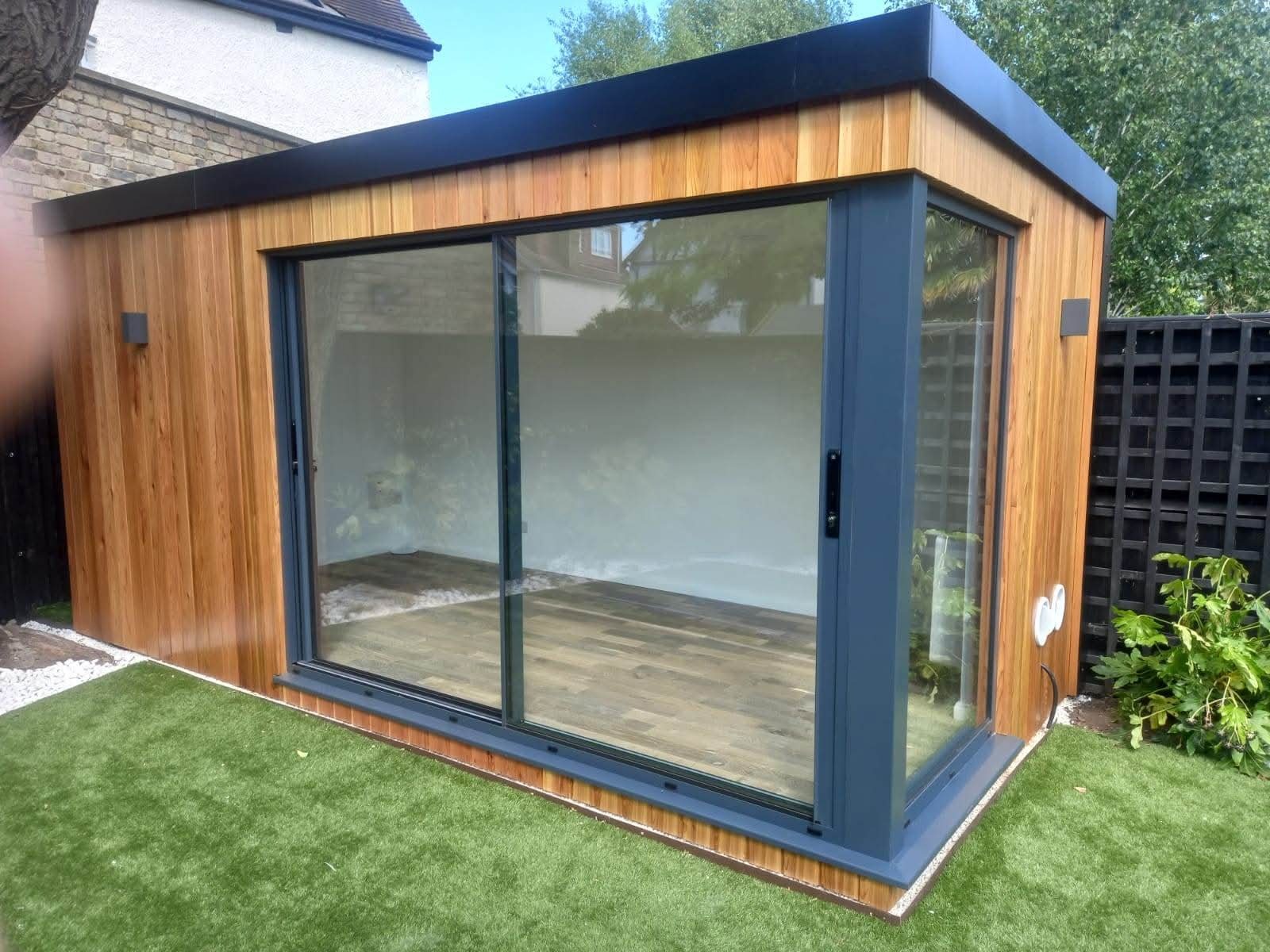 Nowoczesny, drewniany domek ogrodowy z przesuwnymi szklanymi drzwiami, otoczony zielonym trawnikiem i czarnym ogrodzeniem. Widoczne detale wykończenia i oświetlenie zewnętrzne.