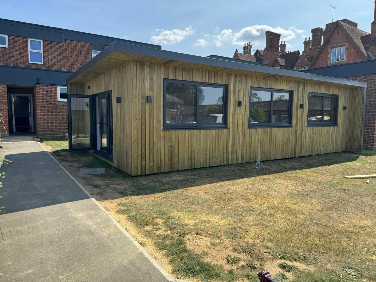 Nowoczesny, parterowy dom modułowy z drewnianą elewacją i dużymi oknami, otoczony trawnikiem. Widoczne drzwi wejściowe i fragment budynku w tle.