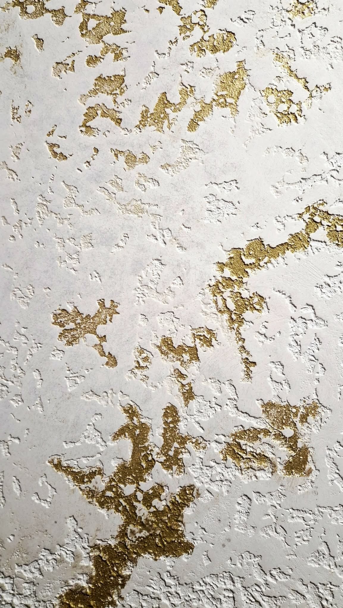 Biała ściana z dekoracyjną, nieregularną fakturą i złotymi akcentami, tworzącymi unikalny wzór. Widoczne nierówności i detale wykończenia.