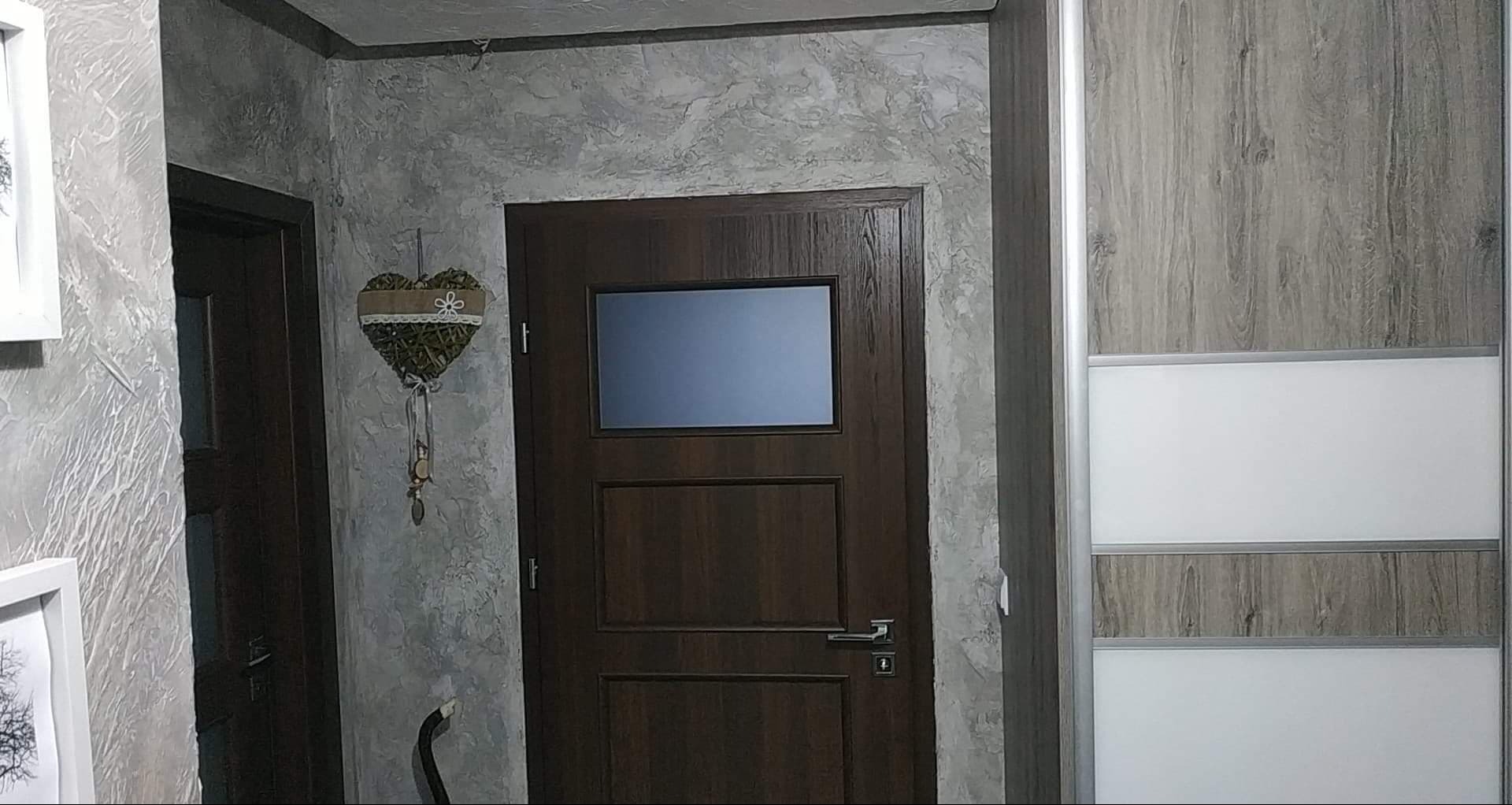 Wykończone wnętrze: dekoracyjna ściana z efektem betonu, ciemne drzwi z szybą i szafa przesuwna z drewnopodobnym wzorem i białymi panelami.