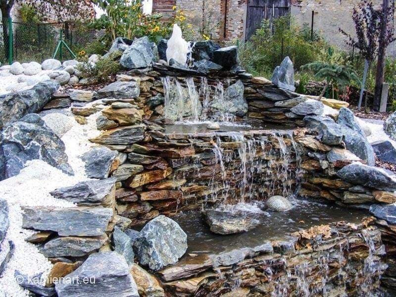 Ogród skalny z kaskadą wodną, zbudowany z płaskich, nieregularnych kamieni w odcieniach szarości i brązu. Woda spływa po kamieniach, tworząc małe wodospady.