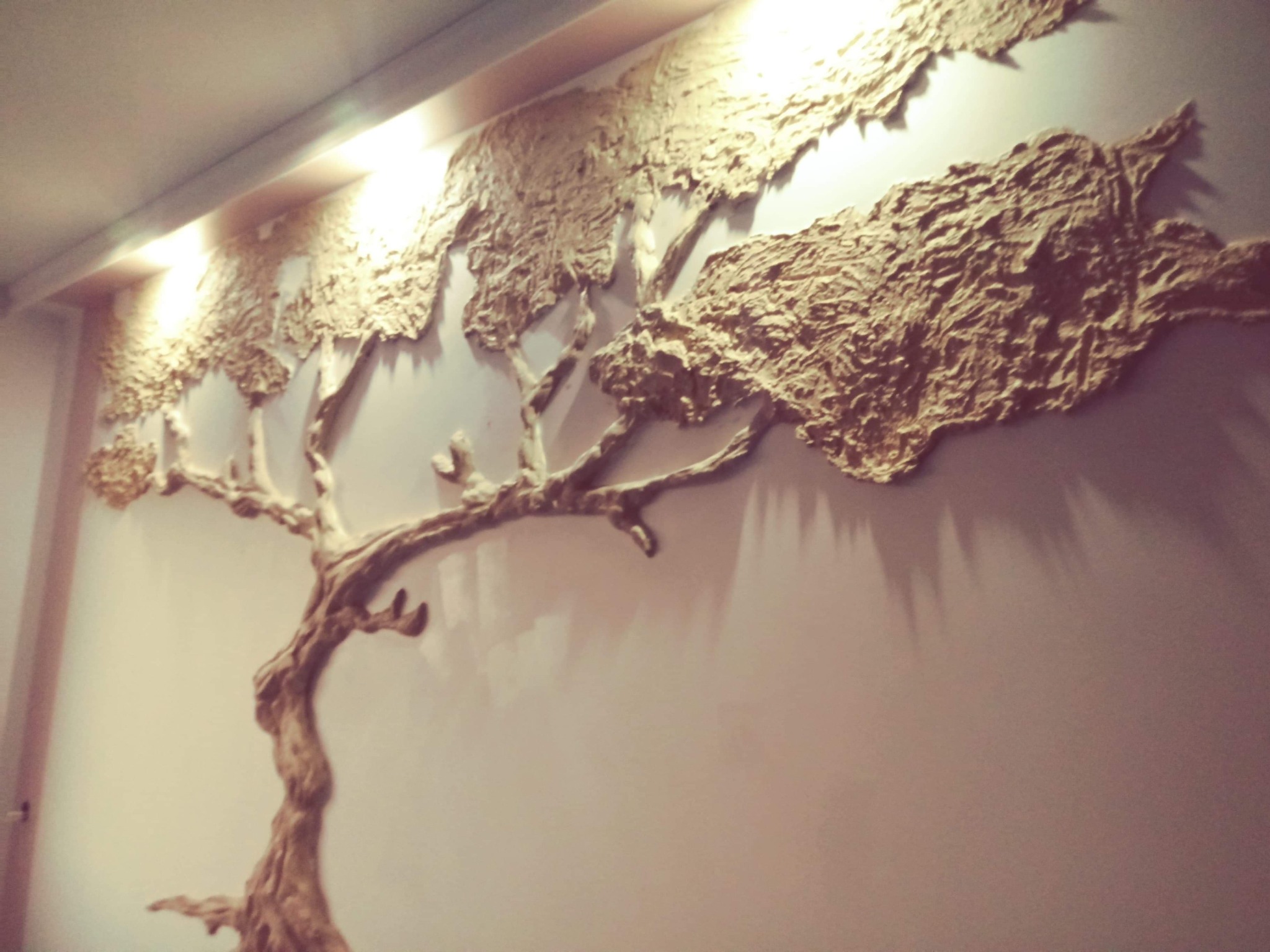Dekoracyjna płaskorzeźba drzewa z fakturą kory na ścianie, oświetlona od góry, tworząca unikalny element wystroju wnętrza.