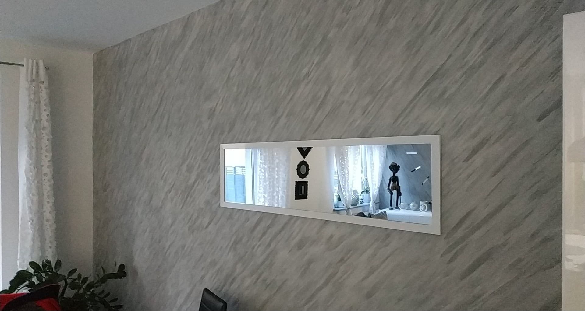 Ściana z dekoracyjną szarą fakturą, na której wisi prostokątne lustro w białej ramie, odbijające okno i figurkę. Widoczny fragment rośliny doniczkowej.