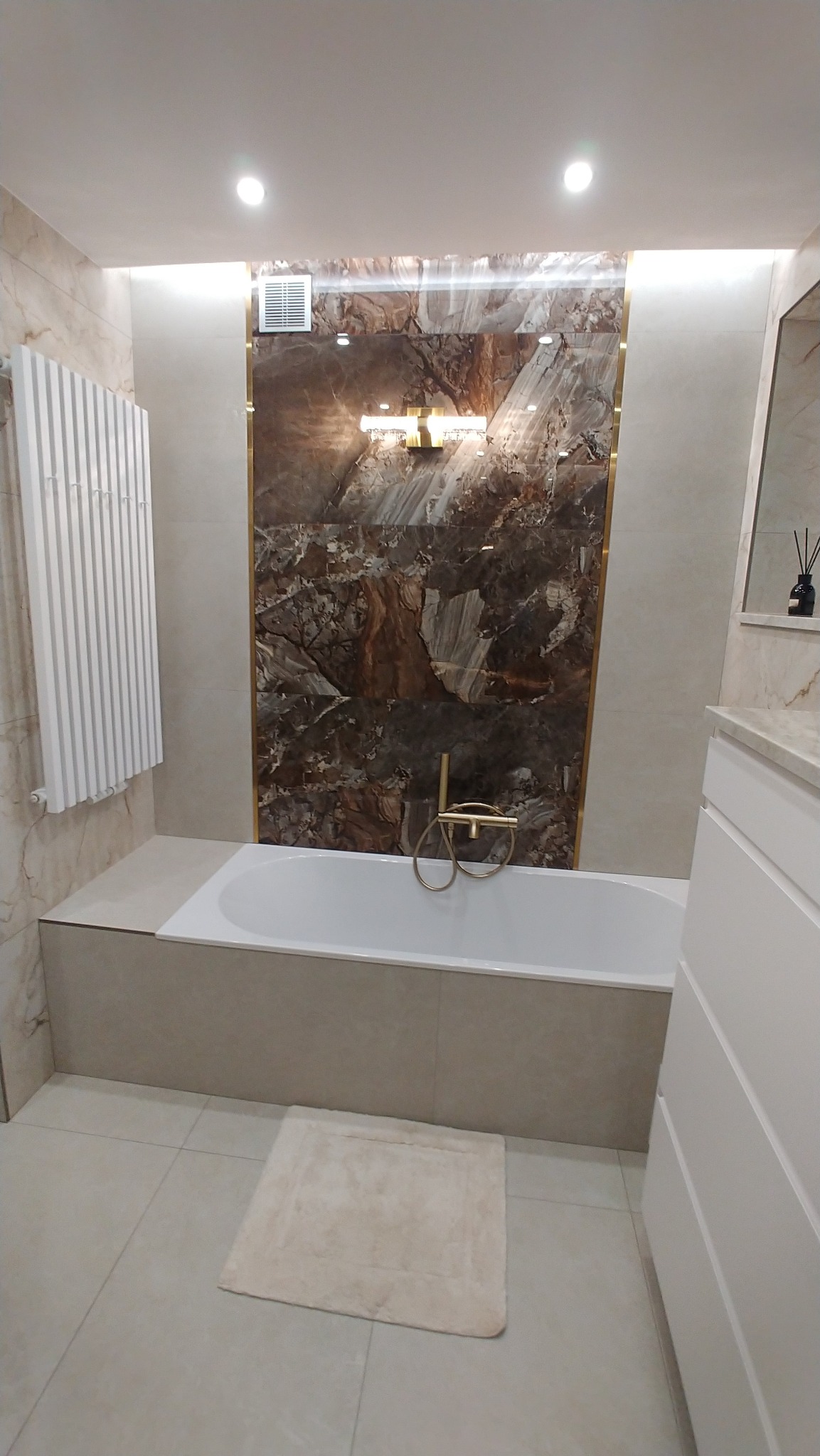 Elegancka łazienka z wanną, marmurową ścianą akcentową w odcieniach brązu i beżu, złotą armaturą i grzejnikiem drabinkowym. Minimalistyczny design z dywanikiem.
