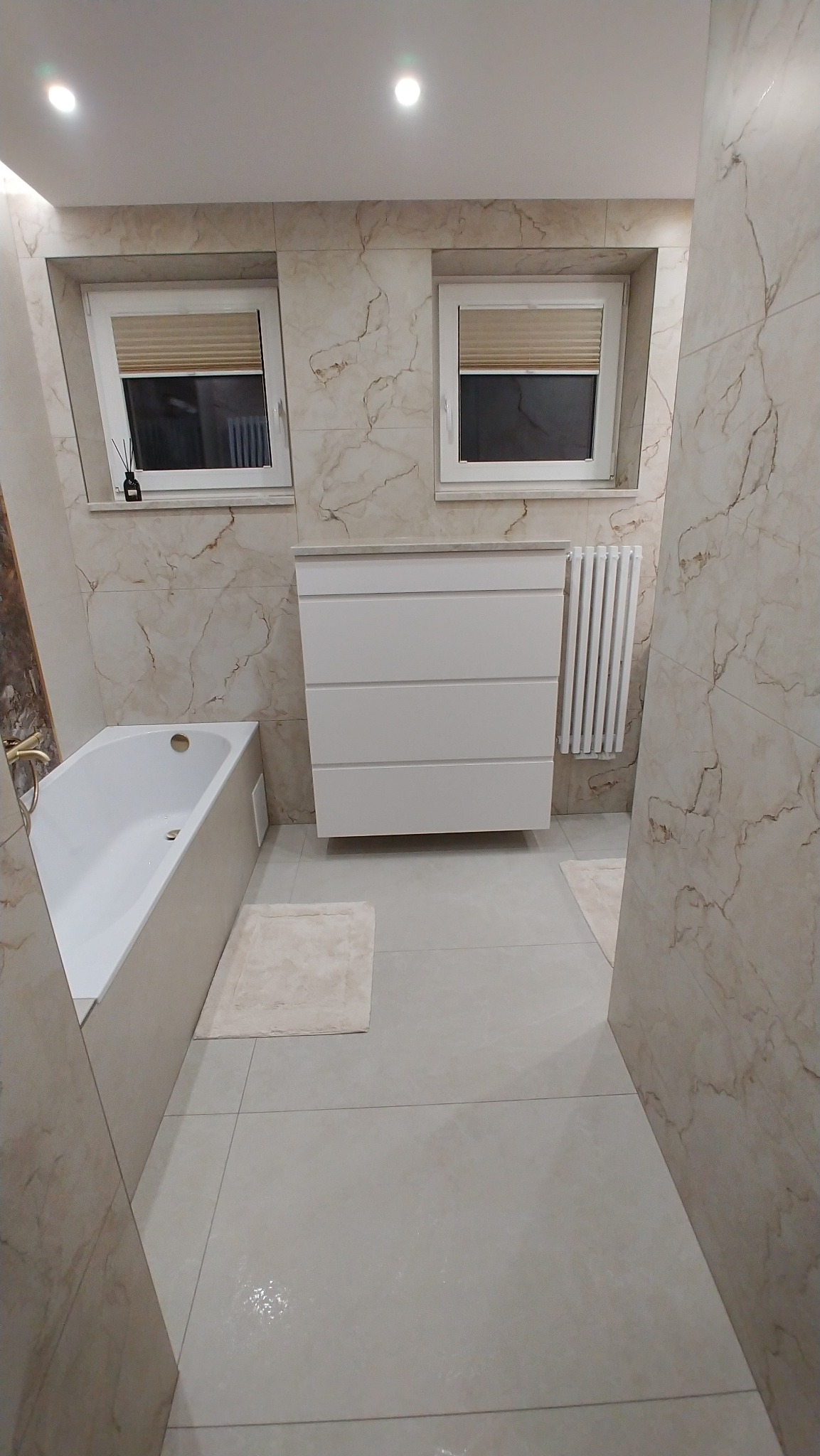 Jasna łazienka z wanną, białą szafką pod umywalką i grzejnikiem. Płytki imitujące marmur na ścianach i podłodze. Dwa okna z żaluzjami.