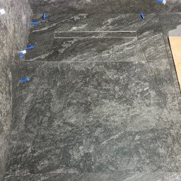 Sebastian Deruś - Ułożone, szare płytki imitujące kamień w kabinie prysznicowej z odpływem liniowym. Widoczne niebieskie krzyżyki dystansowe. Częściowo widoczna płyta z boku.