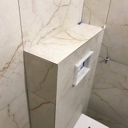 Sebastian Deruś - Obudowa stelaża WC z płytek imitujących marmur, z widocznym otworem na przycisk spłukujący. Użyto listew wykończeniowych. Częściowo widoczna miska WC.