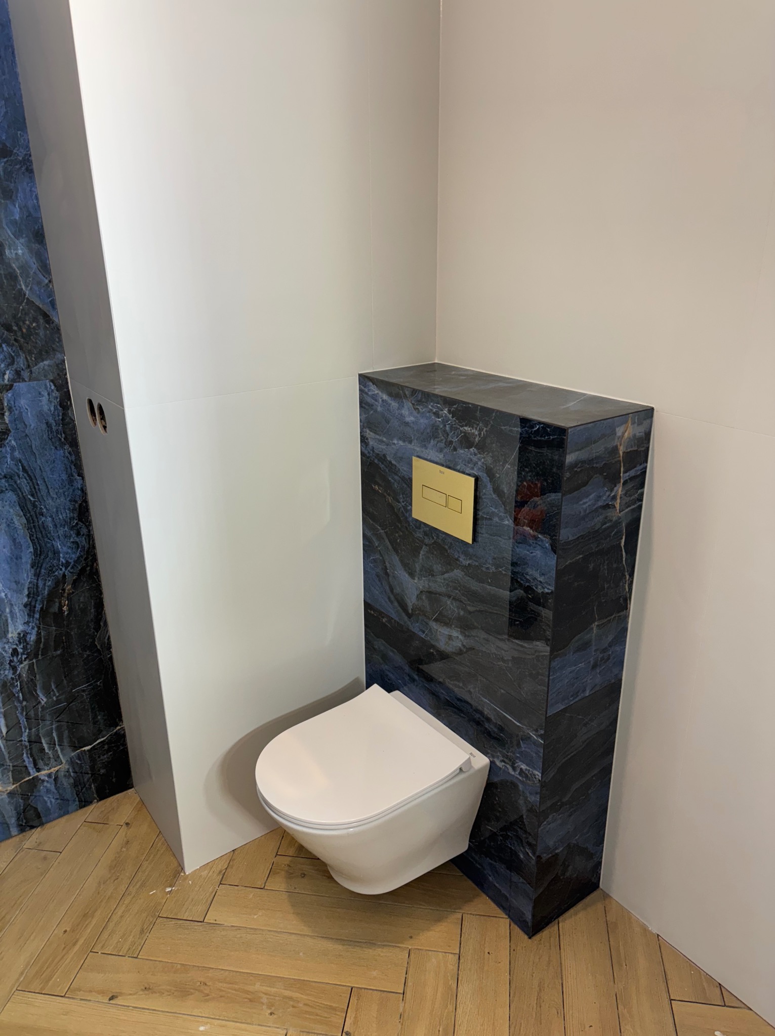 Wykończona łazienka: podwieszana toaleta z marmurową obudową i złotym przyciskiem spłukiwania, drewniana podłoga w jodełkę, białe ściany i zabudowa.