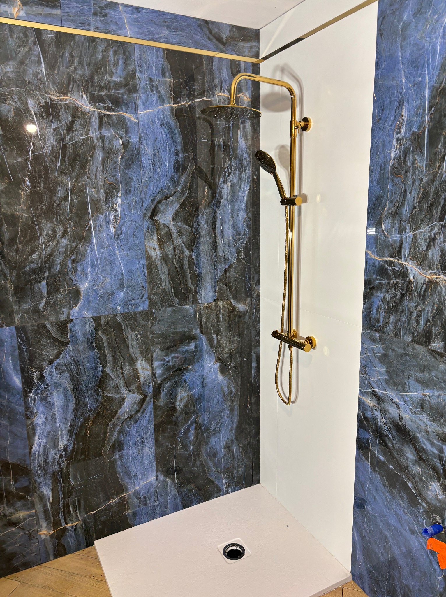 Wykończona kabina prysznicowa z marmurowymi płytkami w odcieniach granatu i złota armatura. Biały brodzik i panel ścienny. Nowoczesny design łazienki.